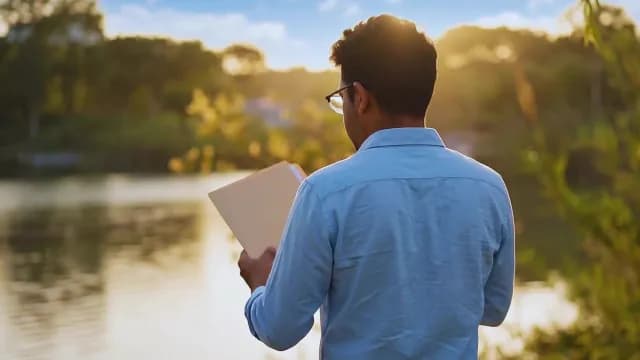 Young Man Reading Book by Peaceful Lake at Sunset