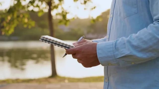 Person Holding Spiral Notebook and Pen Outdoors Near Lake at Sunset